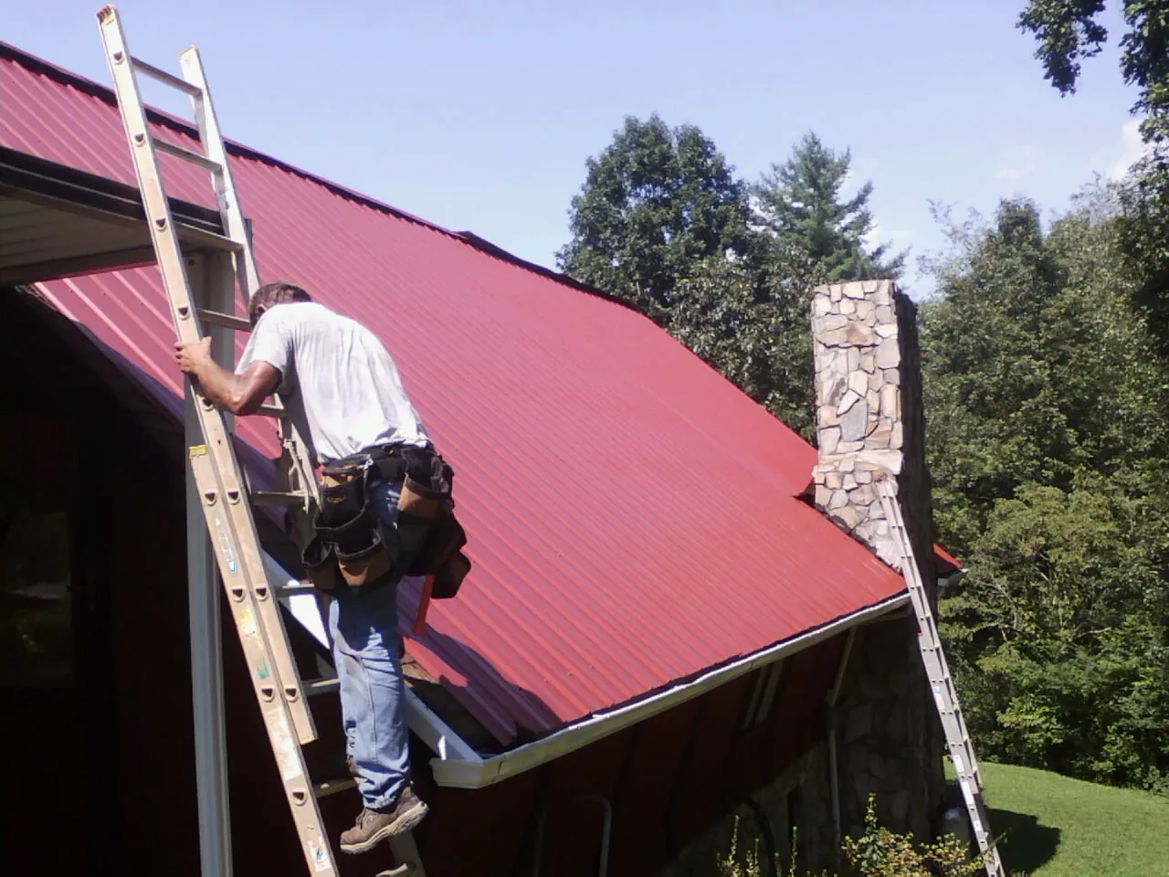 Roofer on a ladder beside a red metal roof and stone chimney — Soffit & Fascia Repair by Roofing Brothers in Ona